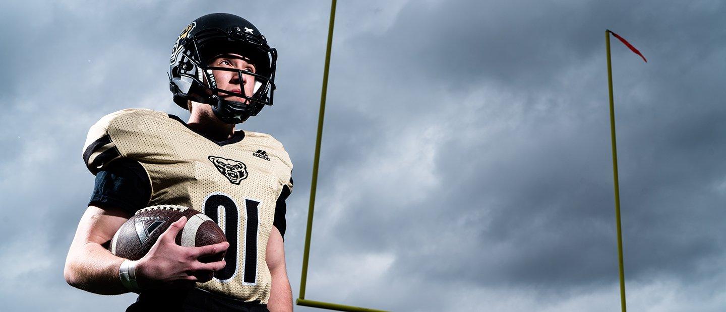 A man in an Oakland University jersey and helmet, holding a football.